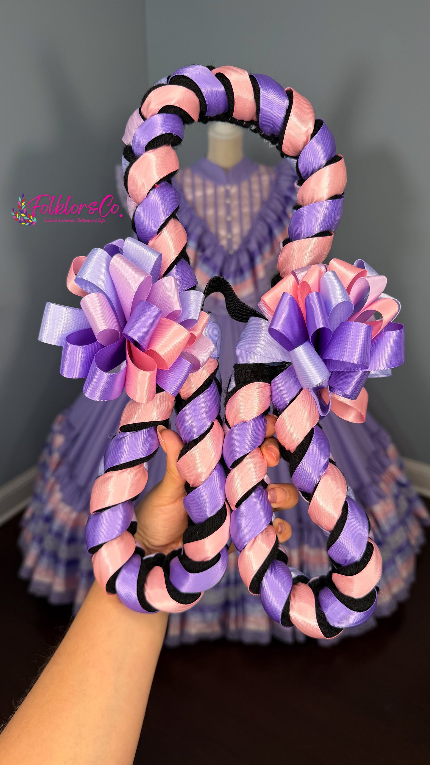 Decorative folklorico trenza with pink and purple ribbons held by a hand against a blurred background